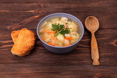 Geleneksel tavuk çorbası, erişte, sebze ve maydanoz yaprakları, iki dilim fırınlanmış ekmek ve ahşap arka planda servis edilir.