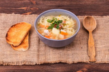 Erişteli tavuk çorbası, sebzeler ve maydanozlu yapraklar. Çuval bezi desenli, açı manzaralı bir tost dilimiyle servis ediliyor.