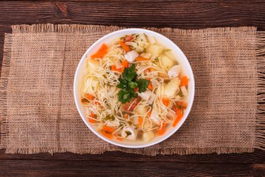 Geleneksel tavuk çorbası, erişte, sebze ve maydanoz yapraklı, dokulu bir çuval bezi, ahşap arka planda, üst görünümde.