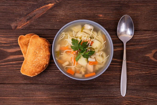 Makarna ve sebzeli ev yapımı tavuk çorbası, bir kaşık ve ahşap masada iki dilim tost.