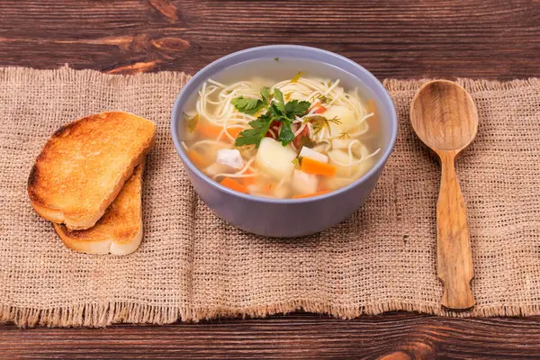 Erişteli tavuk çorbası, sebzeler ve maydanozlu yapraklar. Çuval bezi desenli, açı manzaralı bir tost dilimiyle servis ediliyor.