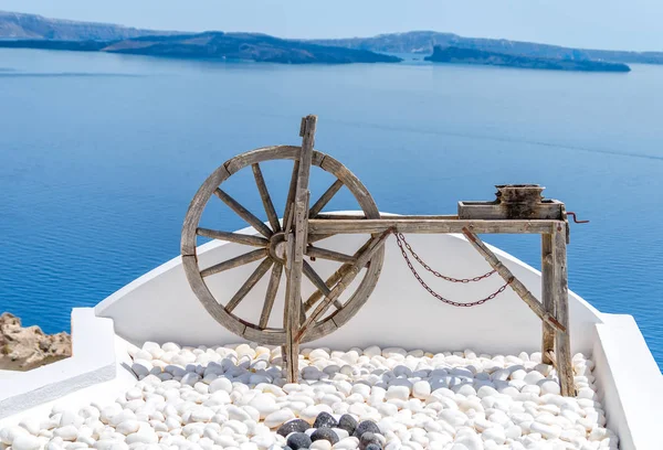 Wooden old tool spinning wheel as a decoration on white stones ground and blue sea rocky landscape in Oia, Santorini island in Greece — Stock Photo, Image