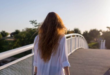 Kızıl saçlı kız bir yaz öğleden sonra gün batımında bir köprüde yürüyor.