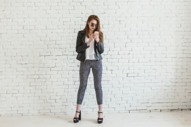 Esmer kız gün ışığında beyaz tuğla duvarın yanında cep telefonu kullanıyor.