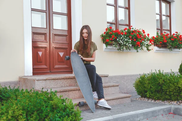 Mujer elegantemente vestida sentada en las escaleras con longboard en las manos delante de la casa . — Foto de Stock