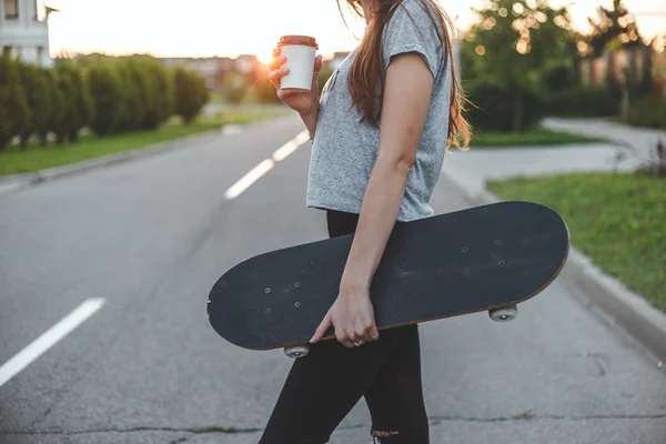 Mooi meisje drinkt uit witte en-klare cup bedrijf skateboard — Stockfoto