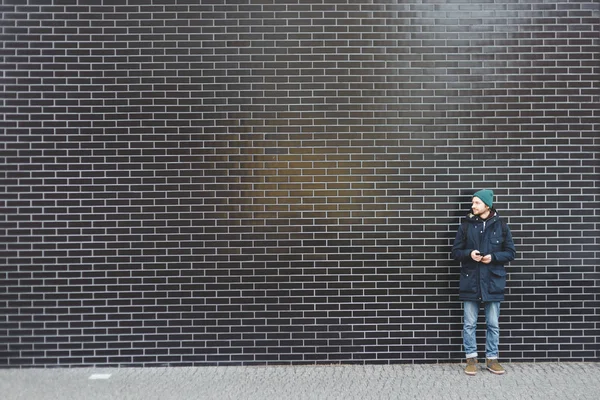 Joven hombre vestido con estilo en ropa de invierno de pie en una pared en blanco de azulejos negros . — Foto de Stock