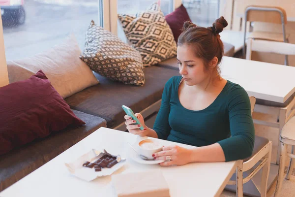 Linda chica leyendo las noticias de la mañana en el teléfono inteligente sentado en la cafetería . —  Fotos de Stock