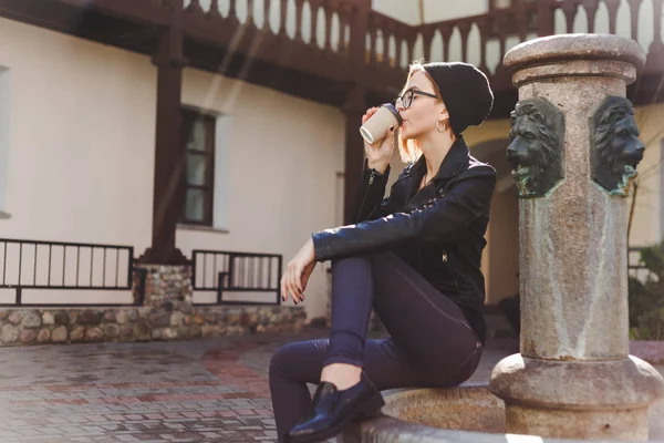 Young woman in black knitted hat and glasses drinking coffee from paper take away cup. — Stock Photo, Image