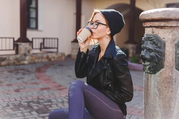 Joven mujer elegantemente vestida en sombrero de punto negro y vasos disfrutando del café de la taza de papel . — Foto de Stock