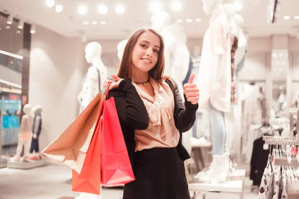 Mladý módní s balíčky plné nákupů ukazuje její postavení palce v butiku — Stock fotografie