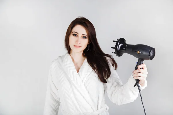 Brunette girl in a bathrobe with a hair dryer dries her hair against a light background — Stock Photo, Image