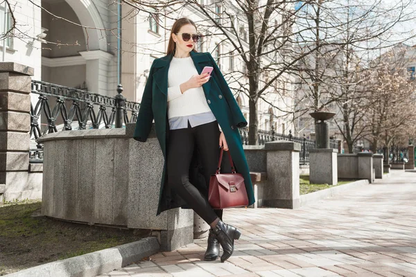 Chica glamurosa vestida con un abrigo caro, un suéter blanco y gafas de sol con un teléfono móvil en la mano posando al aire libre — Foto de Stock