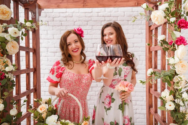 Duas meninas bonitas vestidas em vestidos de verão posando sob um arco de flores com copos de vinho tinto nas mãos — Fotografia de Stock