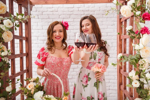 Duas meninas bonitas vestidas em vestidos de verão posando perto de um arco de flor com copos de vinho tinto nas mãos — Fotografia de Stock