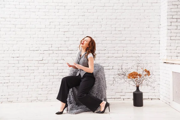 Hermosa mujer está utilizando una tableta digital y sonriendo mientras se sienta en el sillón en el interior moderno —  Fotos de Stock