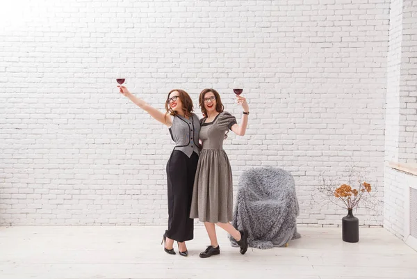 Dos chicas atractivas, divirtiéndose y bebiendo vino tinto en la fiesta. Vestido de moda con hermoso cabello ondulado. En la pared de ladrillo blanco loft —  Fotos de Stock