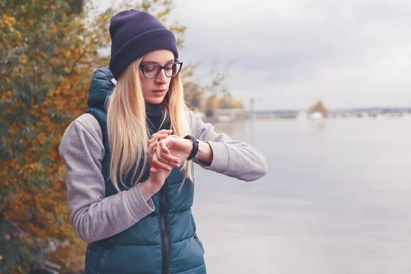 Elegantemente vestida deportista rubia ajusta una pulsera-podómetro electrónico de pie en la orilla del lago. Estilo de deportes de otoño. Viajes y estilo de vida saludable — Foto de Stock