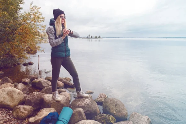 Stijlvol geklede blonde Sportvrouw past een elektronische armband-stappenteller staande op de oever van het meer. Herfst sport stijl. Reizen en gezonde levensstijl concept — Stockfoto