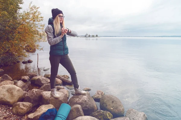 Sportive blonde élégamment habillée ajuste un bracelet-podomètre électronique debout sur la rive du lac. Style sportif d'automne. Concept de voyage et mode de vie sain — Photo