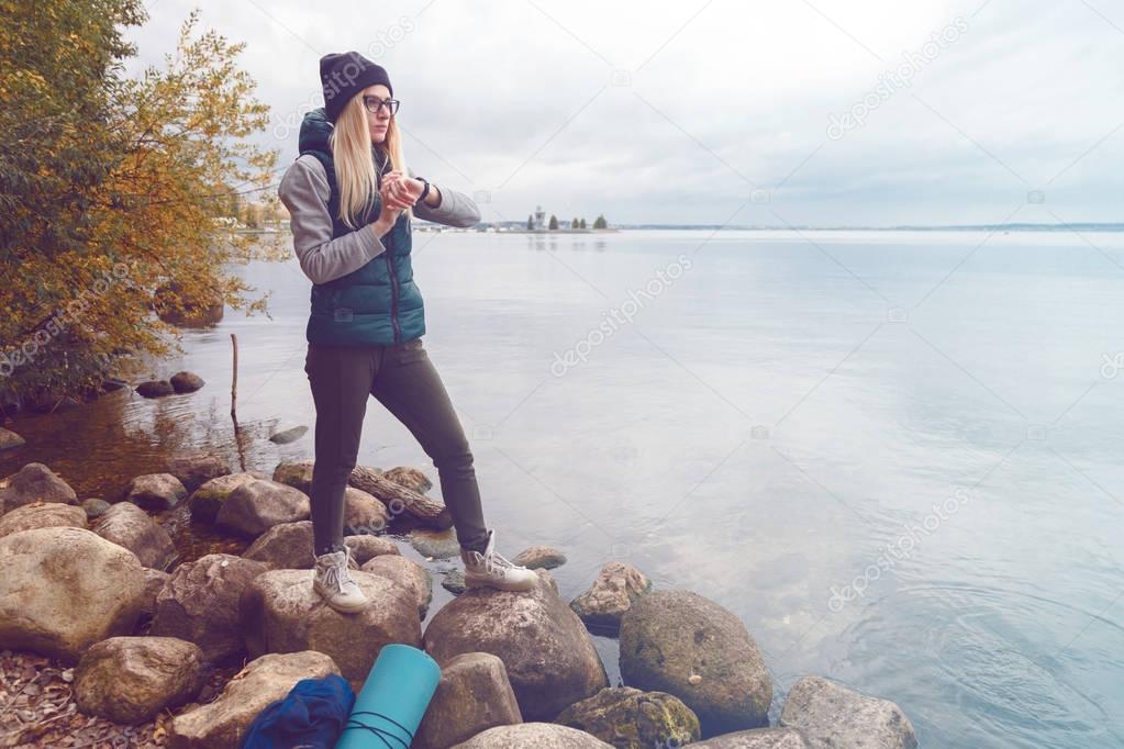 Stylishly dressed blonde sportswoman adjusts an electronic bracelet-pedometer standing on the lake shore. Autumn sports style. Travel and healthy lifestyle concept
