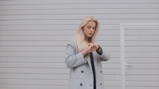 Una joven rubia vestida a la moda con un abrigo usa una pulsera inteligente al aire libre. Vídeo en cámara lenta — Vídeos de Stock