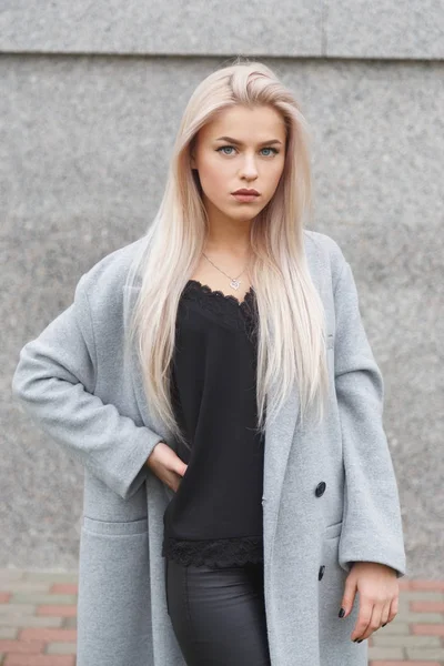 Porträtt av en vacker ung blond kvinna i grå kappa och svart läderbyxor. Street mode look — Stockfoto