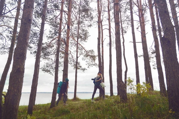 Copines avec sacs à dos marchant à travers la forêt en face du lac et vue sur la forêt. Un voyageur hipster qui explore le monde . — Photo
