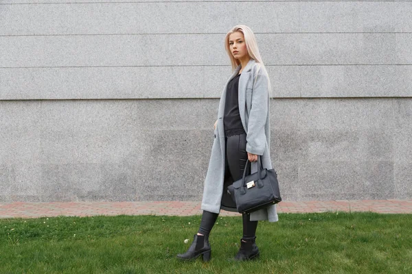 Fashionable woman posing outdoor, holding black leather bag, wearing stylish boots, elegant coat. Female fashion concept. — Stock Photo, Image