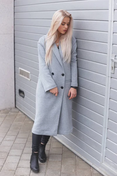 Hipster girl wearing gray coat posing against rough street wall, minimalist urban clothing style — Stock Photo, Image