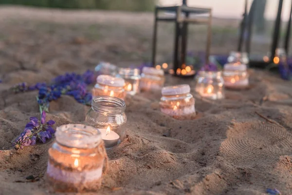 Beautiful decorated romantic place for a date with jars full of candles hanging on tree and standing on a sand. — Stock Photo, Image