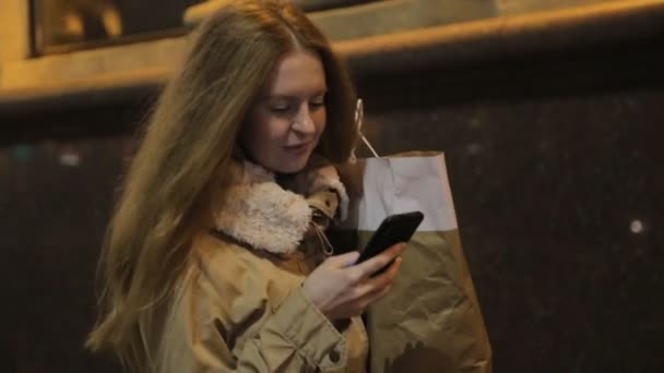 Mujer enamorada de las compras caminando por la noche por la ciudad y mirando el teléfono . — Vídeos de Stock