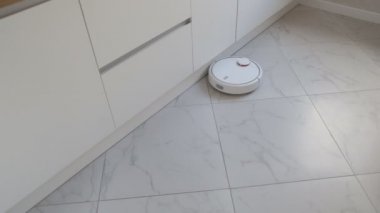 Beyaz robot süpürge mutfak mobilyaları boyunca hareket ediyor.
