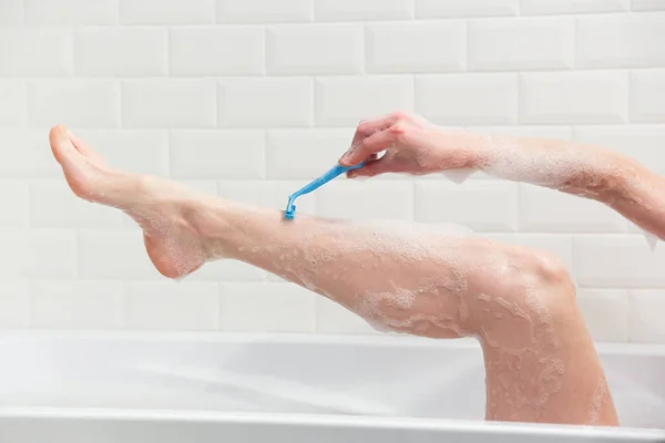 Uma mulher na casa de banho rapa as pernas com uma navalha. Close-up de uma mão com uma navalha . — Fotografia de Stock