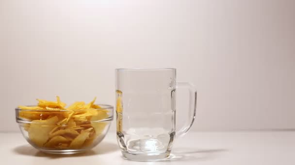 Cerveza vertida de una botella en un vaso de cerveza sobre blanco. Patatas fritas en tazón — Vídeo de stock