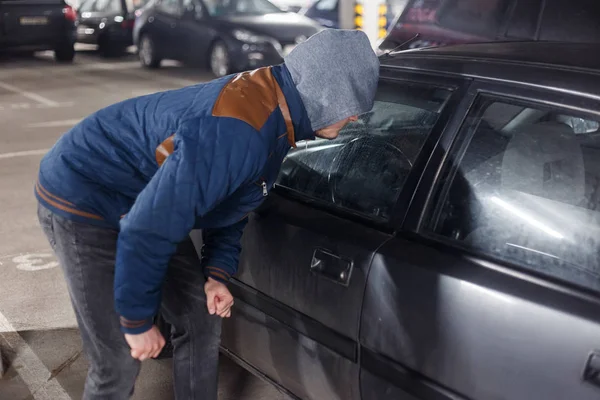 Ladrão de homens de meia preta no rosto e no capô está tentando invadir o automóvel sem sinalizar. O assaltante está a piratear a fechadura da porta do veículo privado. Roubo no conceito de carro. Segurança de estacionamento ruim — Fotografia de Stock