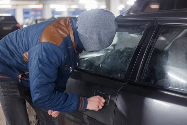 Ladrão de homens de meia preta no rosto e no capô está tentando invadir o automóvel sem sinalizar. O assaltante está a piratear a fechadura da porta do veículo privado. Roubo no conceito de carro. Segurança de estacionamento ruim. — Fotografia de Stock