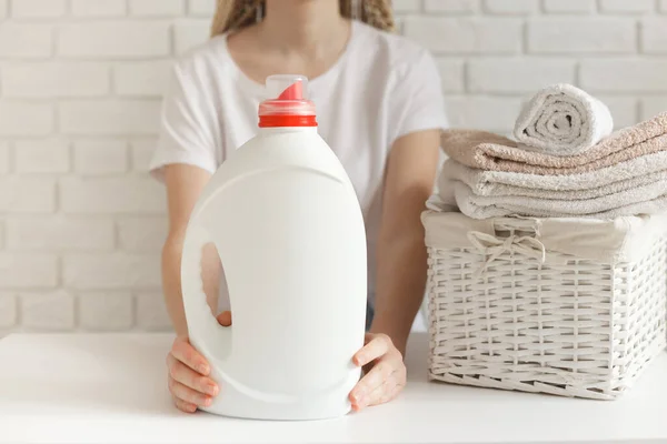 Kobieta trzymająca białą butelkę z detergentem. Wiklinowy kosz z ubraniami i na stole — Zdjęcie stockowe
