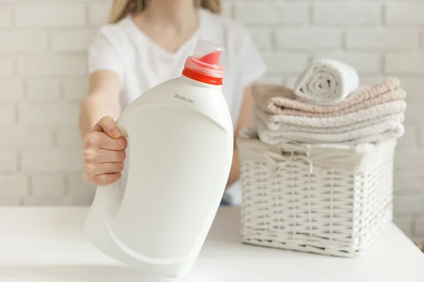 Женщина держит белую бутылку с моющим средством. Плетеная корзина с одеждой и на столе — стоковое фото