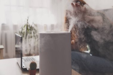 Serbest çalışan bir kadın, iş yerindeki rutubet ve mikroklimayı iş yerinin iş yerinde dizüstü bilgisayar ve belgelerle korumak için ev içi nemlendirici kullanır..