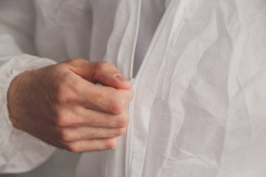 Doktor epidemiyoloji uzmanı beyaz koruyucu giysi giyer, fermuarı bağlar, elleri yakınlaştırır. Elleri koruyucu lastik eldiven içinde. Kişisel koruyucu ekipman konsepti.