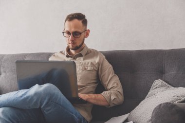 Beyaz, genç, serbest çalışan bir adam. Evden çalışıyor. Kanepede dizüstü bilgisayarla oturmak. İnternet sitelerinde gezinmek ya da çalışmak