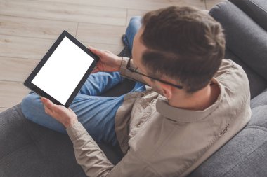 İnternet sörfü ve konferans çağrısı. Adamın biri evdeki kanepede otururken tablet bilgisayar kullanıyor. Günlük giysiler giymişti. Boş ekranlı tablet.