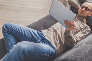 İnternet sörfü ve konferans çağrısı. Adamın biri evdeki kanepede otururken tablet bilgisayar kullanıyor. Günlük giysiler içinde.