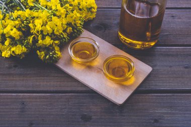 Masanın üzerinde bir şişe ve bir kase kolza tohumu yağı var. Tahta masadaki genç kolza tohumu çiçeklerinin yanında.