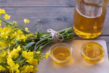 Ağaç tohumu yağıyla dolu iki bardak ve ahşap bir masada taze kolza tohumu çiçeklerinin yanında bir şişe yağ.