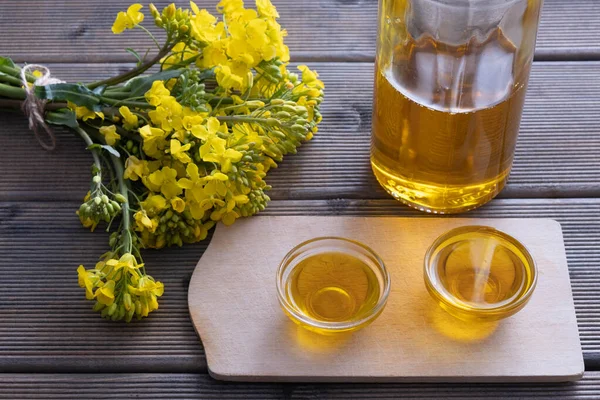 small cups with rapeseed oil and a glass bottle with rapeseed oil next to them are rapeseed flowers on a wooden table