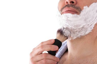 Adam, beyaz arka planda bir fırçayla yanaklarına ve boynuna tıraş köpüğü sürer.
