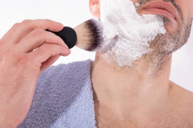 Adam yüzüne beyaz bir arka planda fırçayla tıraş köpüğü sürüyor.