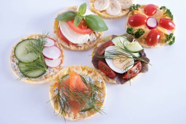 Masanın üzerinde salatalık, yumurta, domates, muz ve turplar beyaz arka planda mısır ekmeğine konmuş.